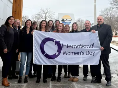 A group of people standing outside a building in winter, holding a white flag that reads “International Women’s Day” with a purple circular symbol. Snow surrounds the area, and the Midland logo is visible in the background.