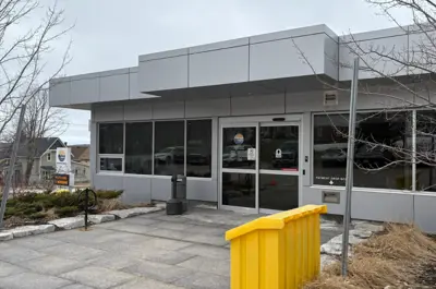 The entrance to the Midland Municipal Office on an overcast day, showing glass double doors, large windows, a paved walkway, and a bright yellow salt storage container in the foreground.