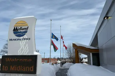 The Midland Municipal Office entrance in winter, with three flags flying at half‑mast beside the walkway. A large roadside sign reads “Welcome to Midland,” and deep snow lines the path under a partly cloudy sky.