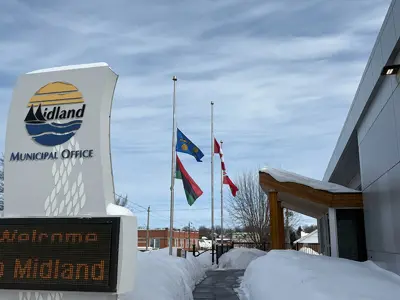 The Midland Municipal Office entrance in winter, with three flags flying at half‑mast beside the walkway. A large roadside sign reads “Welcome to Midland,” and deep snow lines the path under a partly cloudy sky.