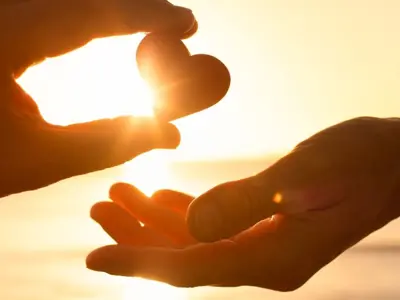 Two hands silhouetted against a golden sunset, with one hand holding a small heart shape and the other reaching toward it. Sunlight shines through the heart, creating a warm glow.